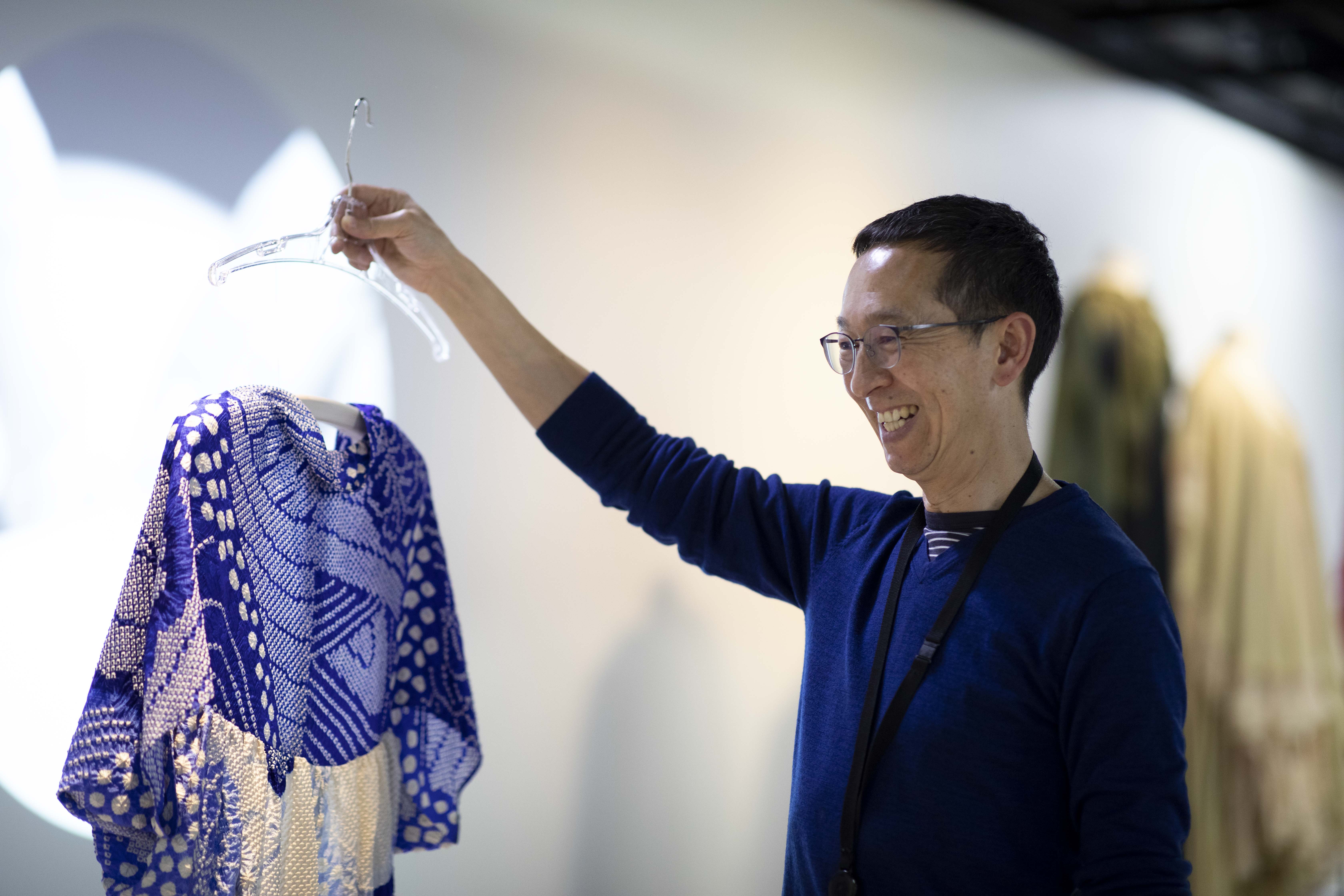 A man holding a coathanger from which drapes a blue and white patterned silk top. The man has a broad smile.